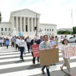 People standing in front of the Supreme Court holding signs and behind a podium