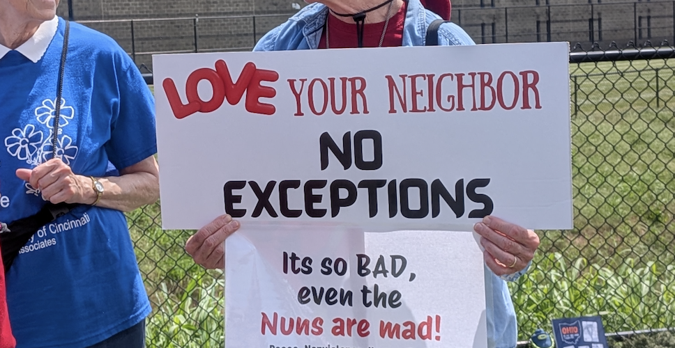 A white woman in sunglasses carries a sign that says "Love your neighbor, no exceptions" and "its so bad even the nuns are mad"